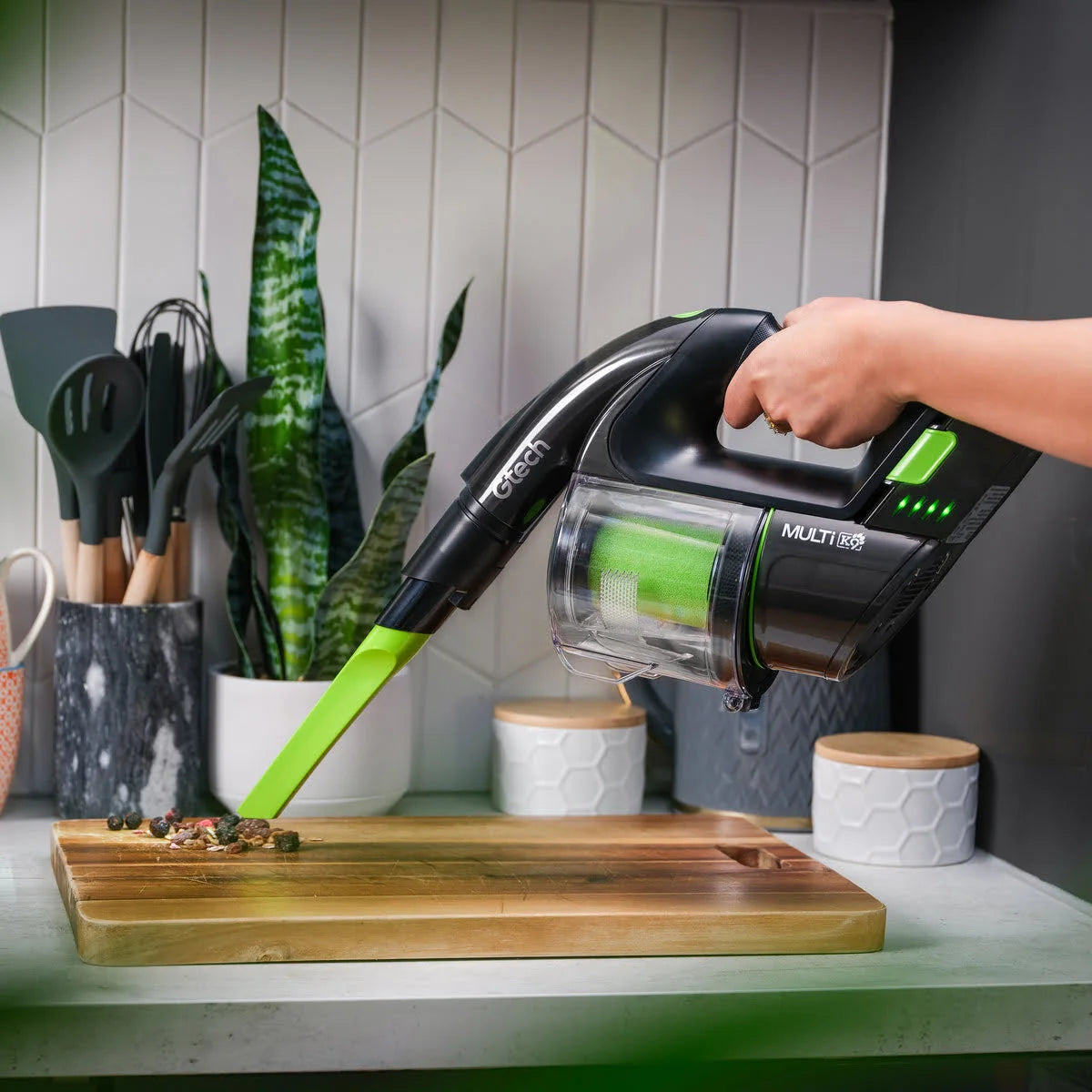 Person using Multi K9 Pet handheld vacuum cleaner on a kitchen counter with plants and utensils in the background.